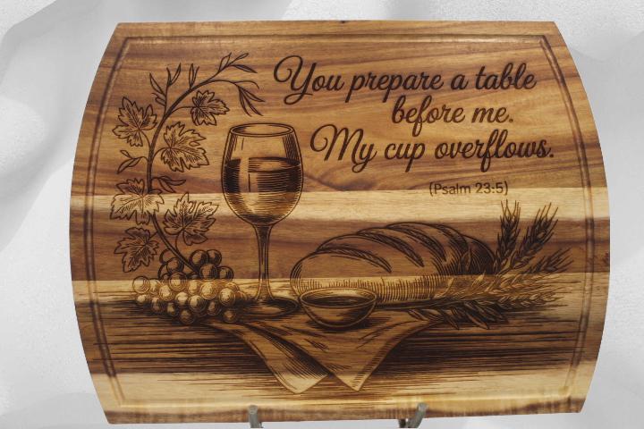 Wooden cutting board with engraved design of a wine glass, bread, and grapes, along with text.
